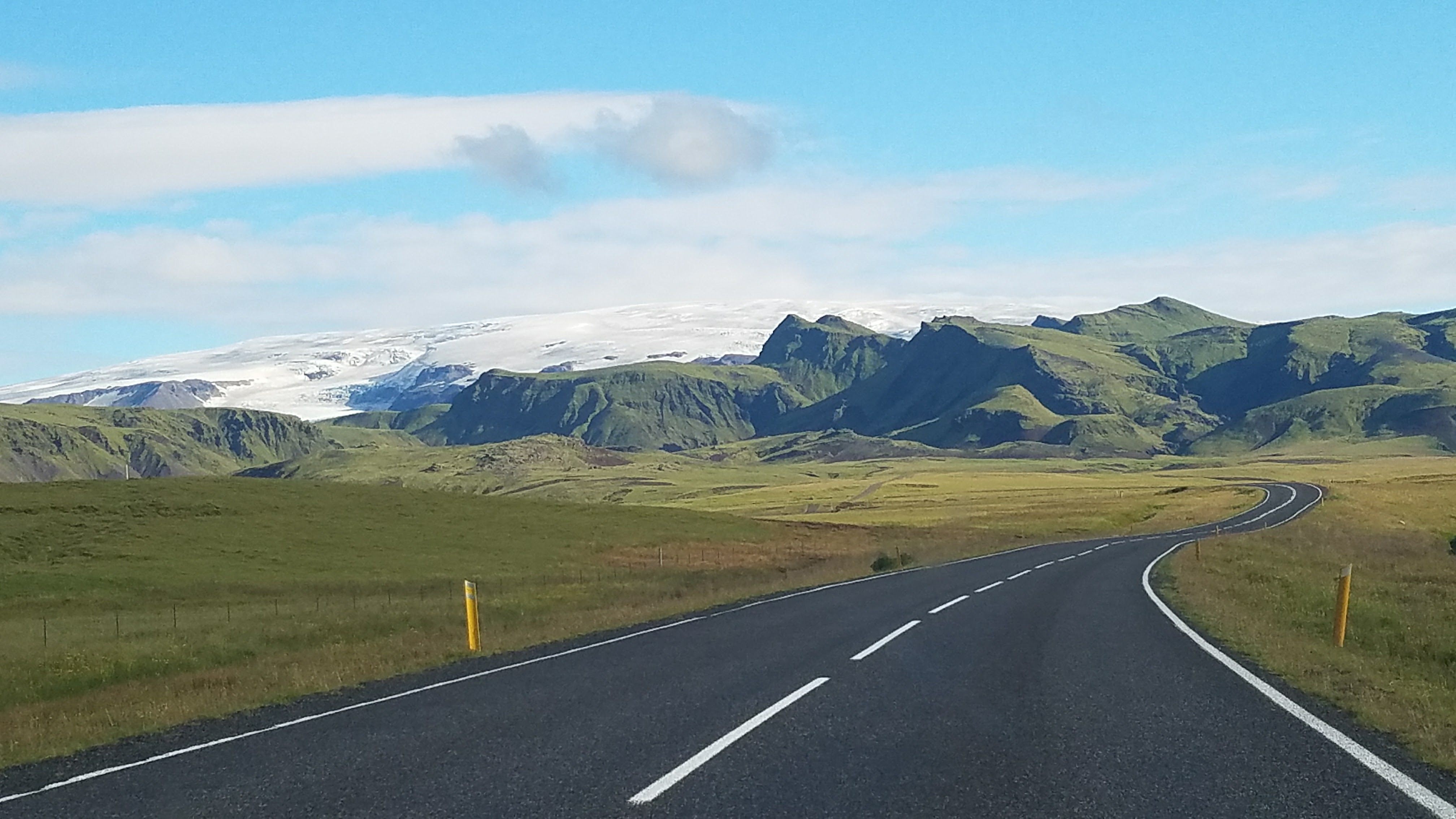 A photograph of a road in Iceland. The road stretched off into rolling green hills. A glacier can be seen in the distance