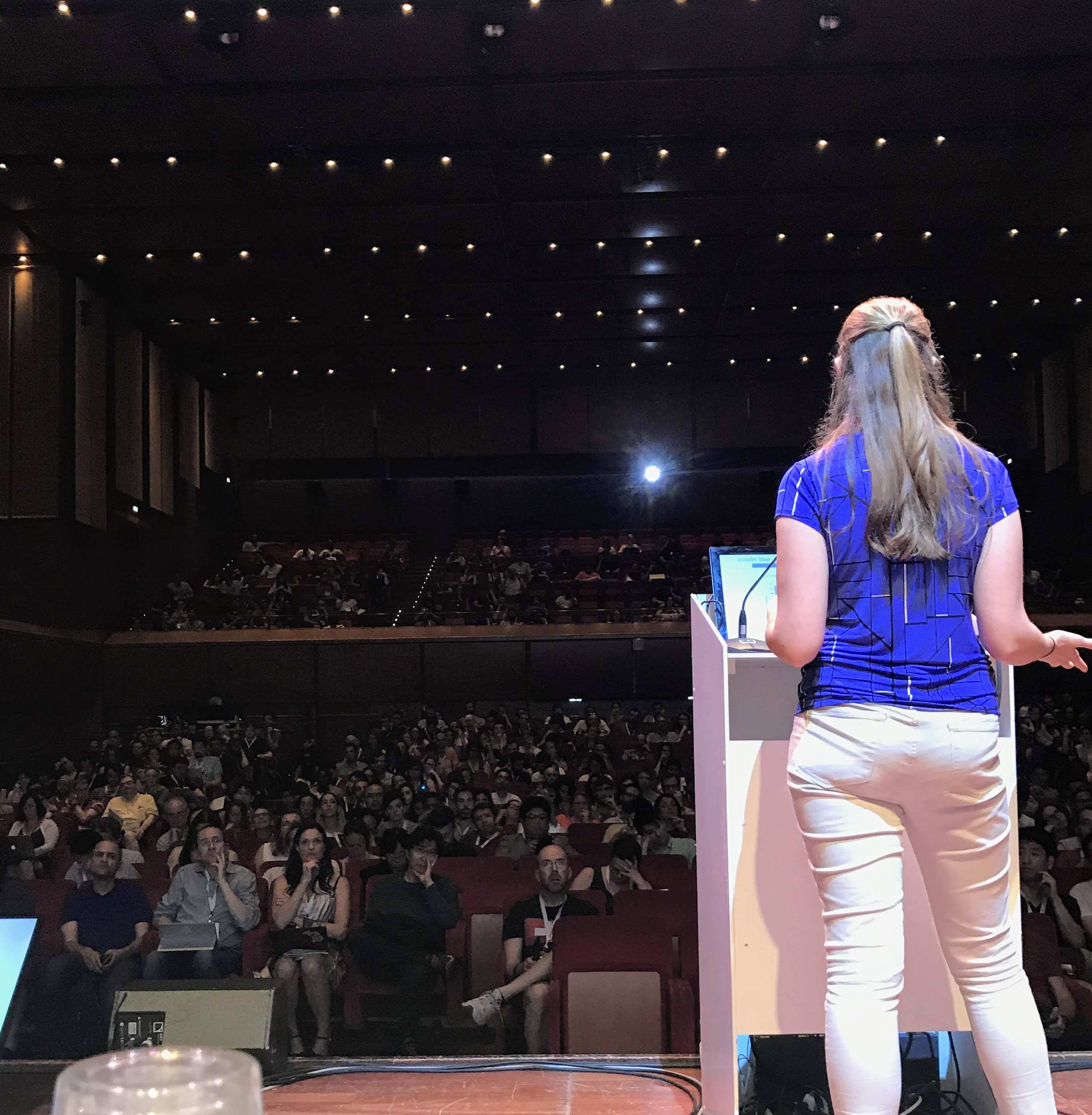 Jennifer is on stage giving a presentation, facing the audience in a large auditorium